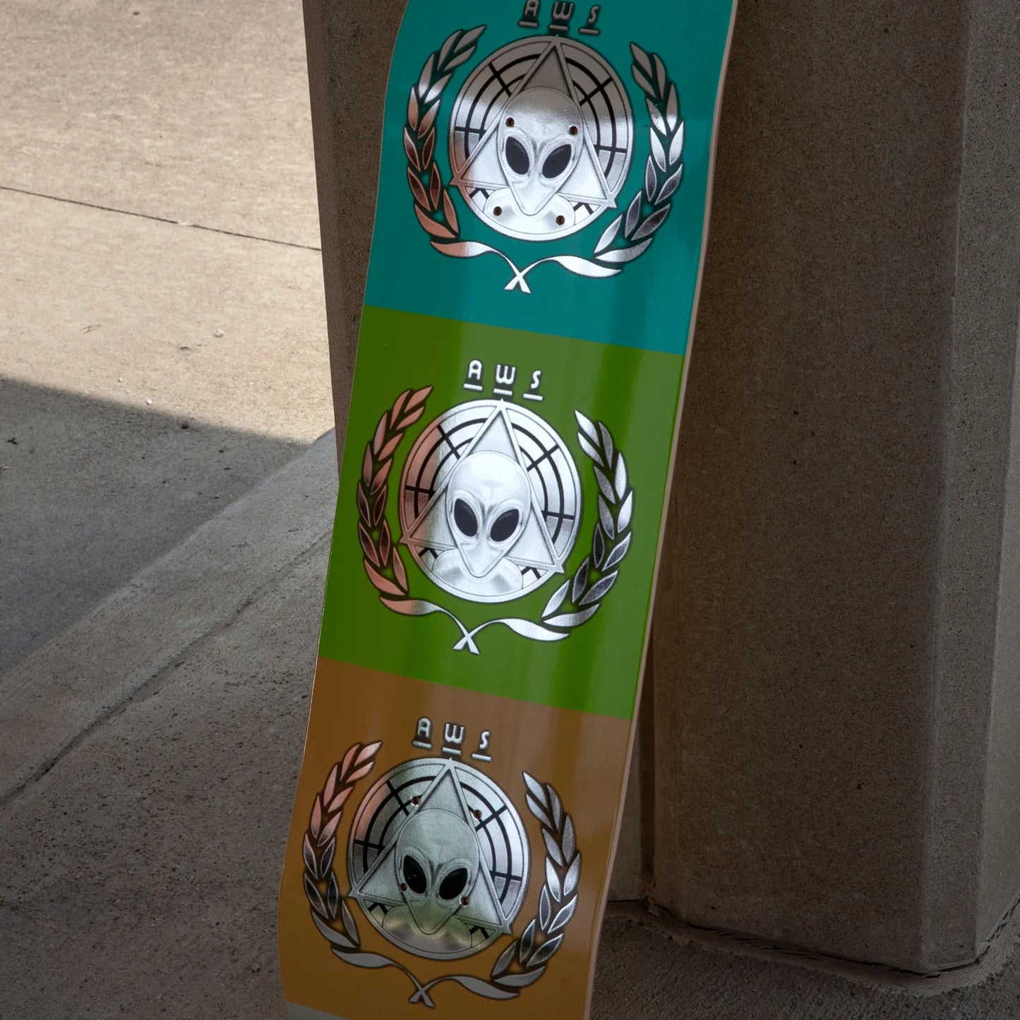 A close-up of a skateboard deck leaning against a concrete post outdoors. The deck features three horizontal color bands: teal, lime green, and tan, each printed with a large silver alien head emblem inside a wreath, with the text 'AWS' above it.