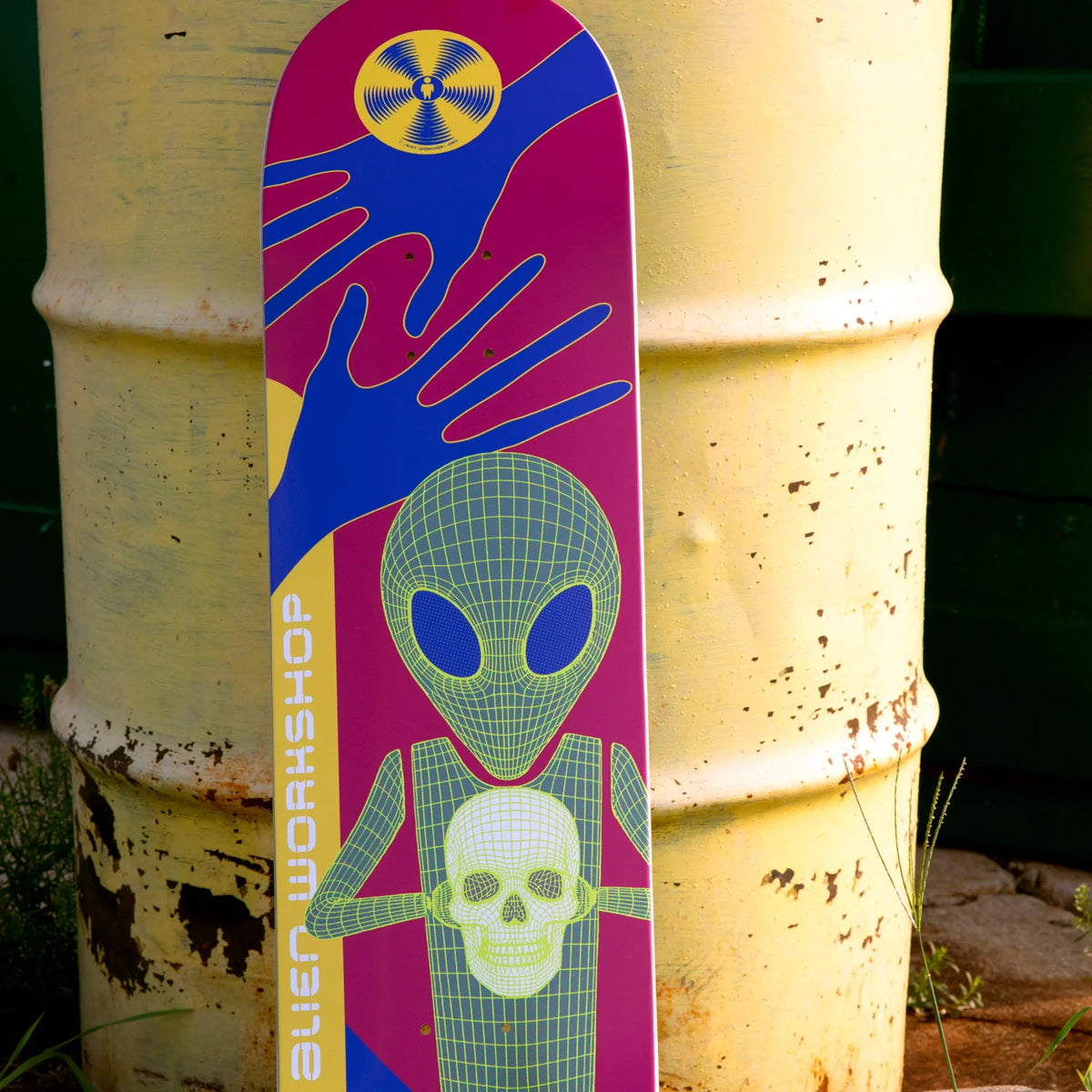 A skateboard deck leaning against a large yellow metal barrel outdoors. The deck features a colorful graphic design with a green wireframe alien holding a skull, blue and magenta abstract shapes, and a yellow circular symbol near the top.