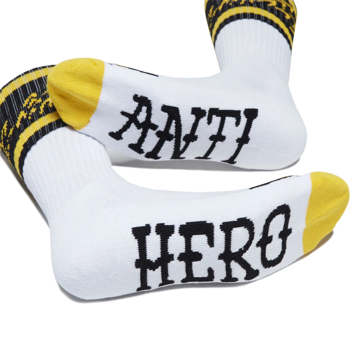 A pair of white crew socks with yellow toes and heels, featuring black text that reads 'ANTI HERO' on the soles, with yellow and black striped cuffs.