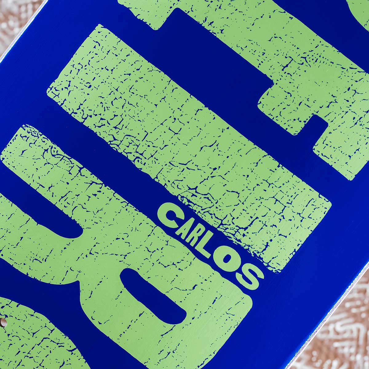Close-up of a skateboard deck featuring a vibrant blue background with large, distressed lime green graphic lettering and the name 'CARLOS' printed in white.