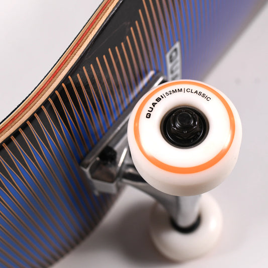 Close-up of a skateboard wheel and truck, showing a white Quasi 52mm classic wheel with an orange ring and black center, attached to a skateboard deck with blue and black graphics.