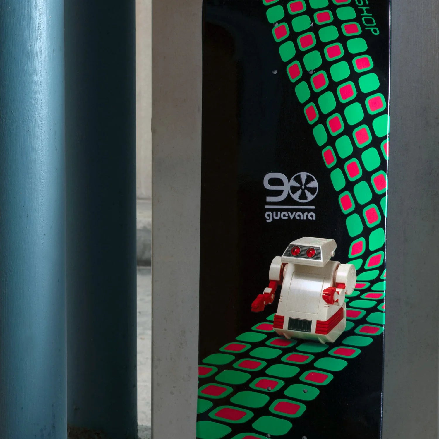 A small white and red toy robot stands on a black printed panel with a green and pink geometric pattern. The panel has the text '90 guavara' and 'shop' visible, and the photo is framed by gray vertical posts on both sides.