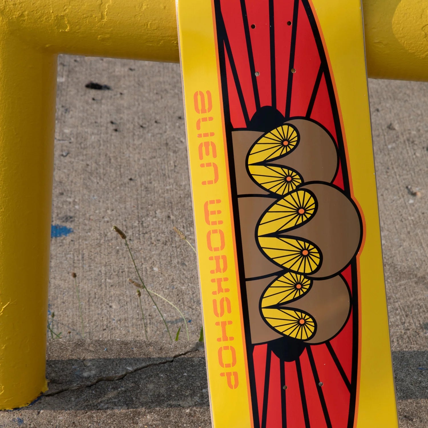 A close-up of a skateboard deck featuring a bold graphic design with a yellow background, red sunburst elements, and a stylized yellow and brown eye motif. The words 'alien WORKSHOP' are printed vertically in orange-red on the side.