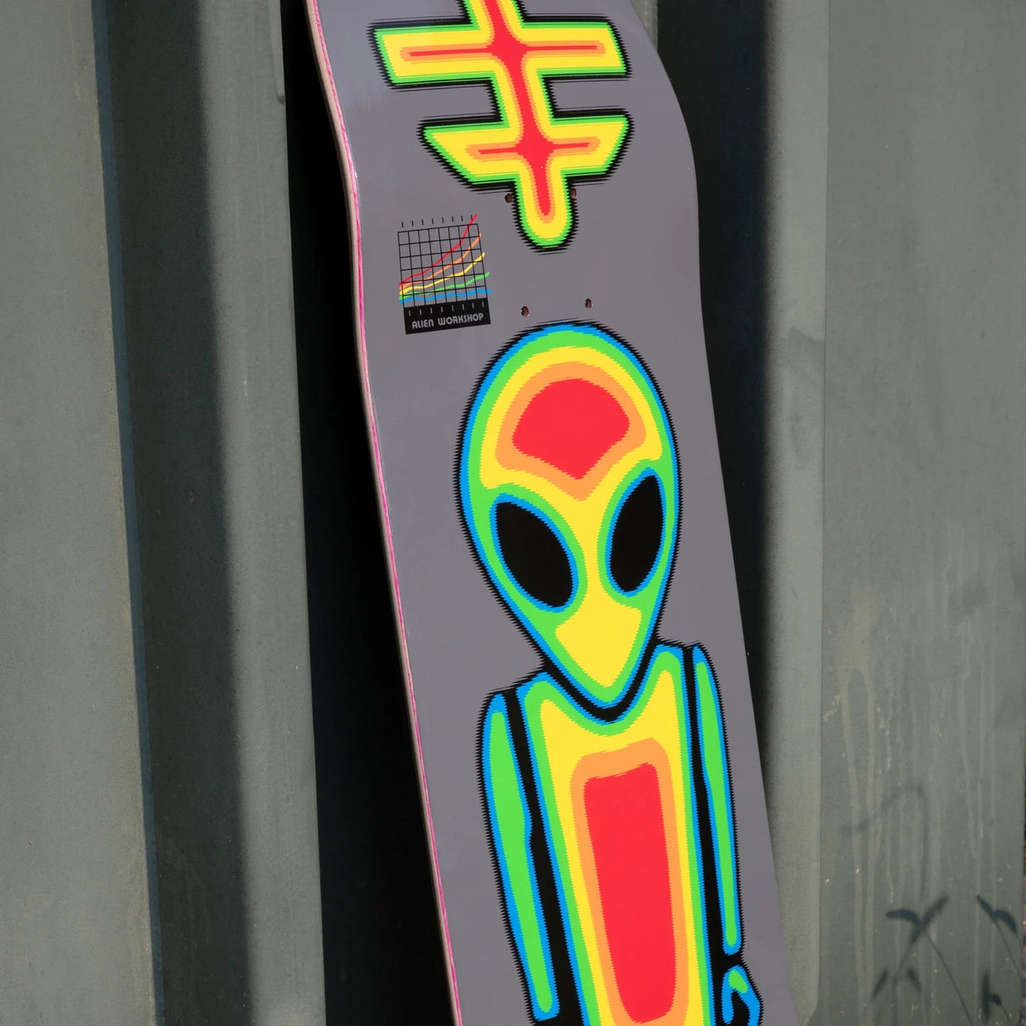 A skateboard deck leaning against a dark gray wall, featuring a large neon-style alien graphic in yellow, green, blue, and red on a dark gray background, with a small sticker near the top.
