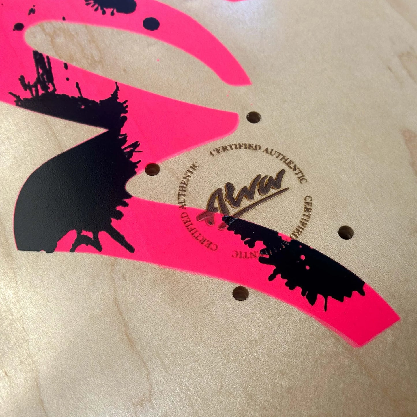 Close-up of a skateboard with pink and black design and 'Certified Authentic' logo.