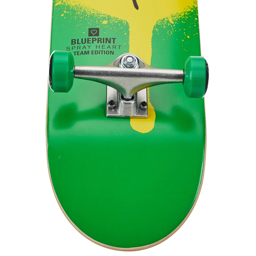 A close-up of a skateboard deck featuring a bright green and yellow gradient design with a black spray-painted heart logo and 'BLUEPRINT SPRAY HEART TEAM EDITION' text, shown with green wheels and silver trucks against a white background.