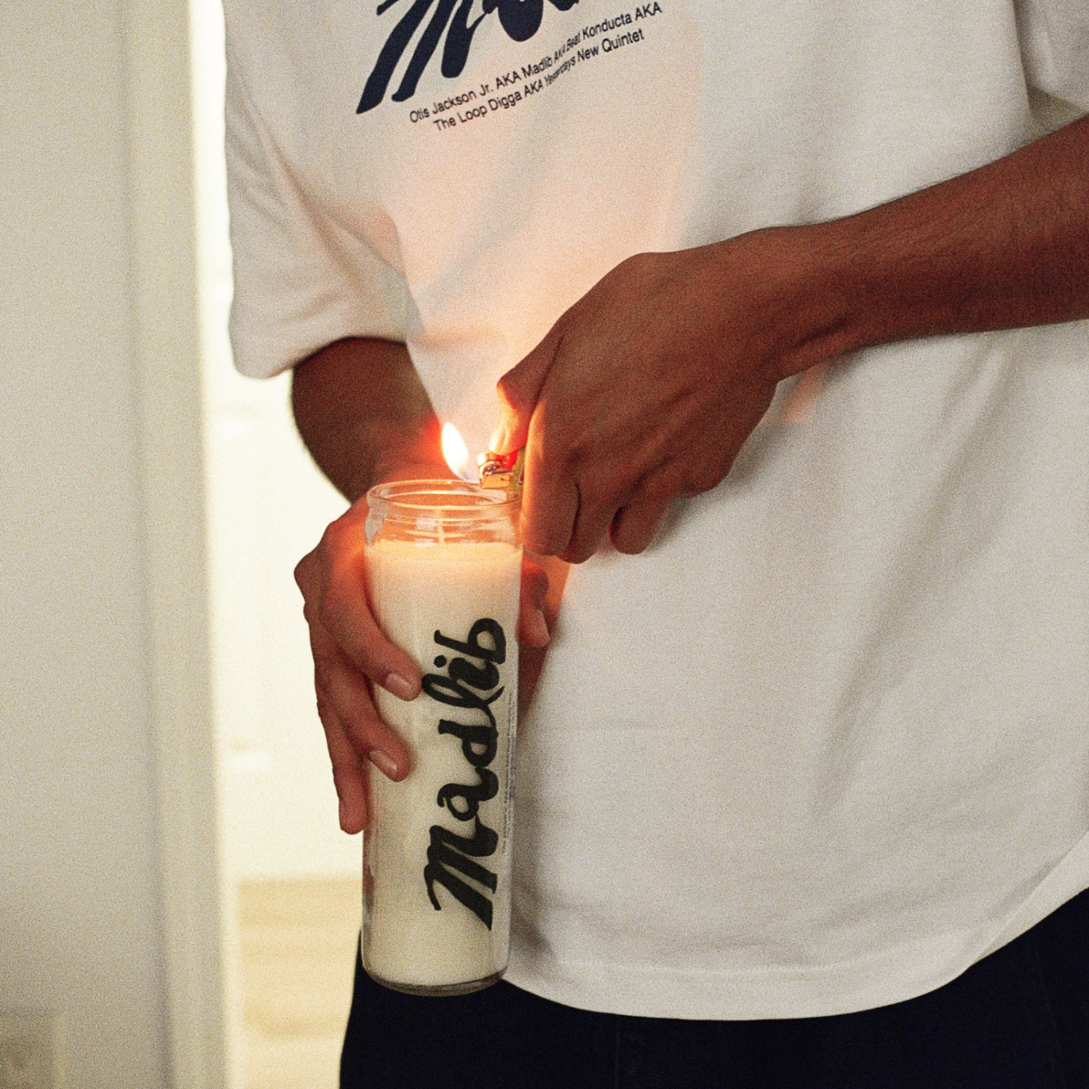 Person holding a candle with 'Madlib' branding, wearing a white t-shirt.