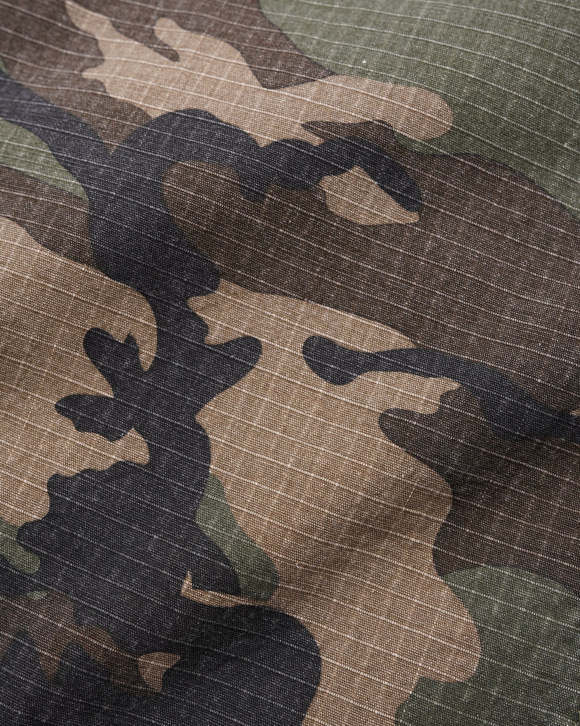 Close-up of a camouflage fabric with a subtle grid texture, featuring organic shapes in dark brown, tan, olive green, and black.