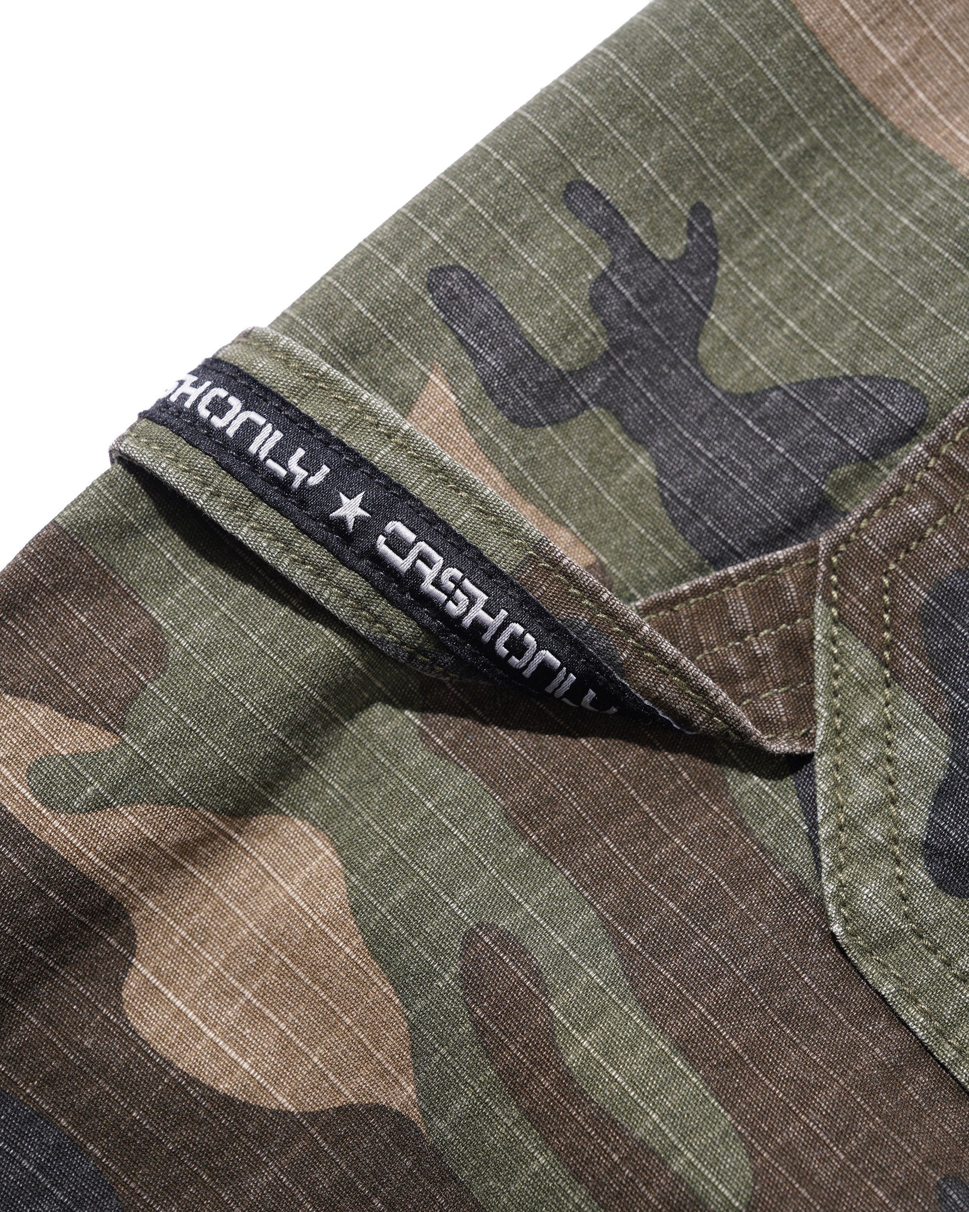 Close-up of a camouflage-patterned garment, likely a shirt or jacket, showing a collar with a black fabric brand label that reads 'CASHONLY' and 'CASHONLY'.