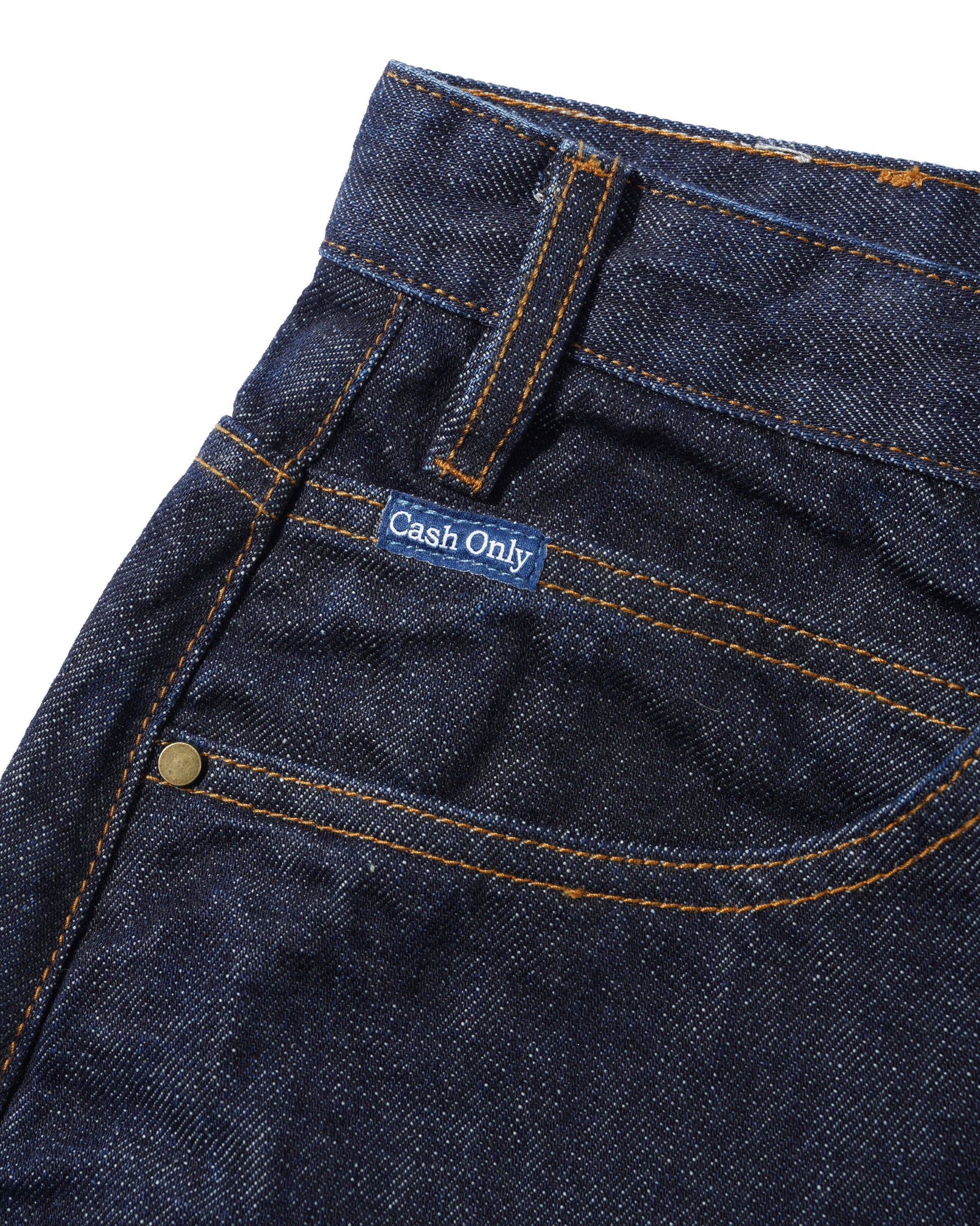 Close-up of dark blue denim jeans showing the waistband, belt loop, front pocket, and a small blue label that reads 'Cash Only'.