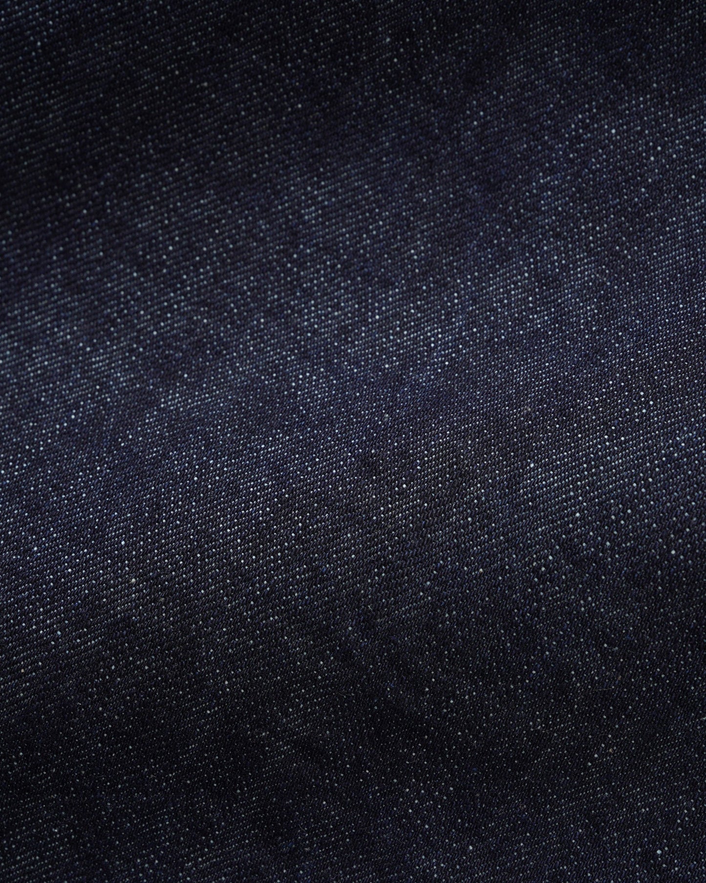 A close-up texture shot of dark indigo denim fabric, showing the fine twill weave and subtle color variation.