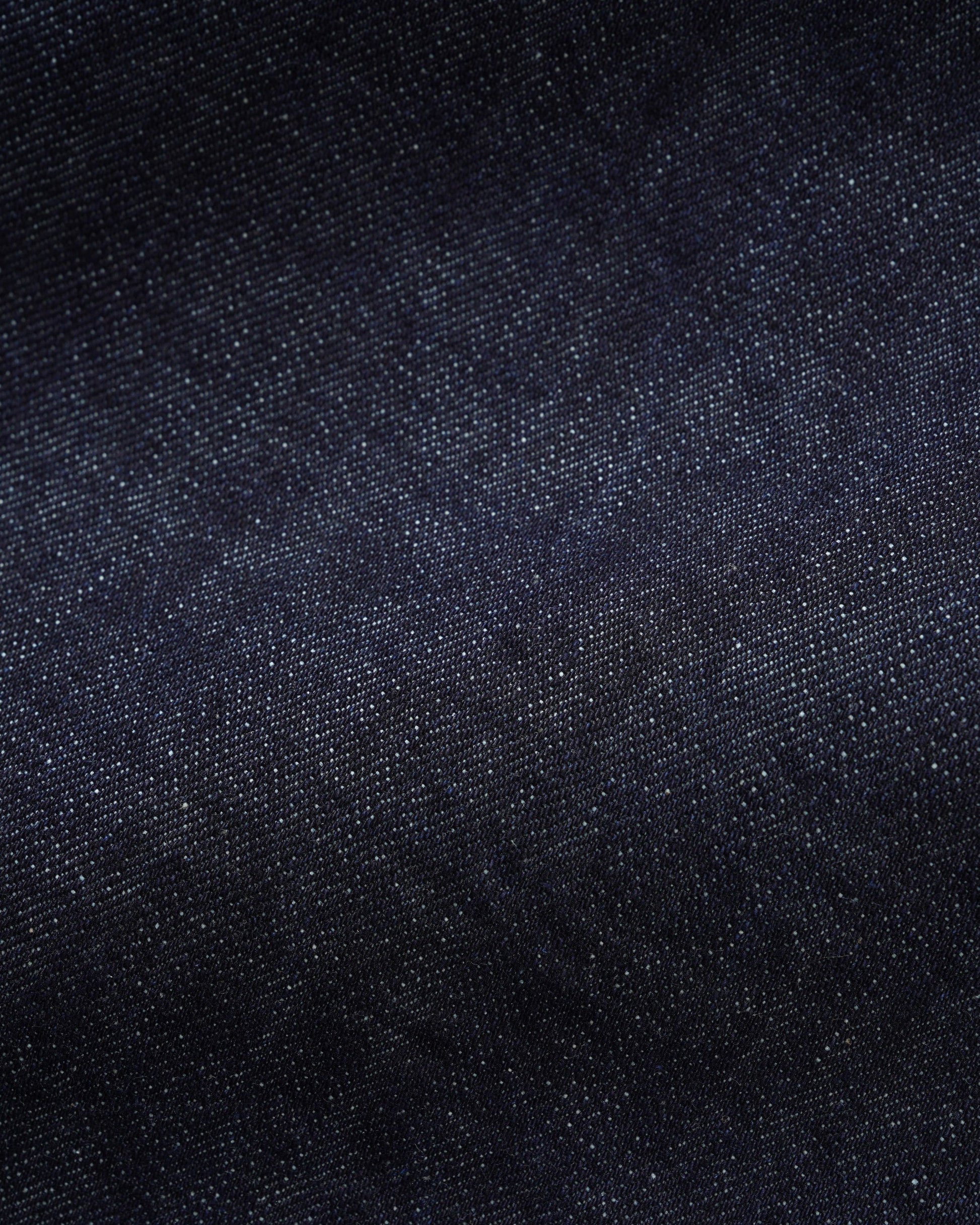 A close-up texture shot of dark indigo denim fabric, showing the fine twill weave and subtle color variation.