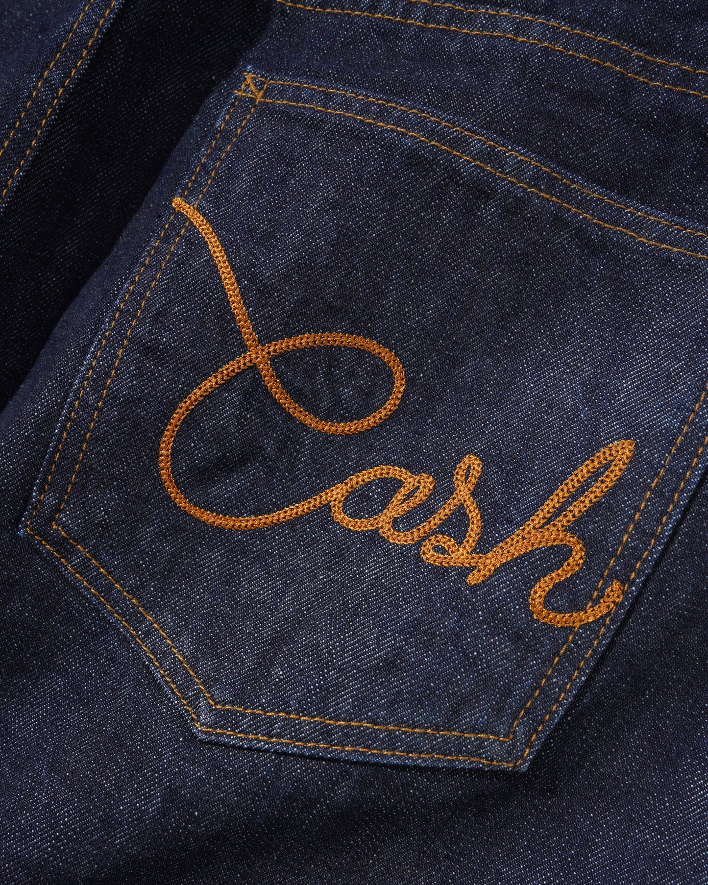 Close-up of a dark indigo denim pocket with orange embroidered script reading 'Cash' and matching orange contrast stitching.
