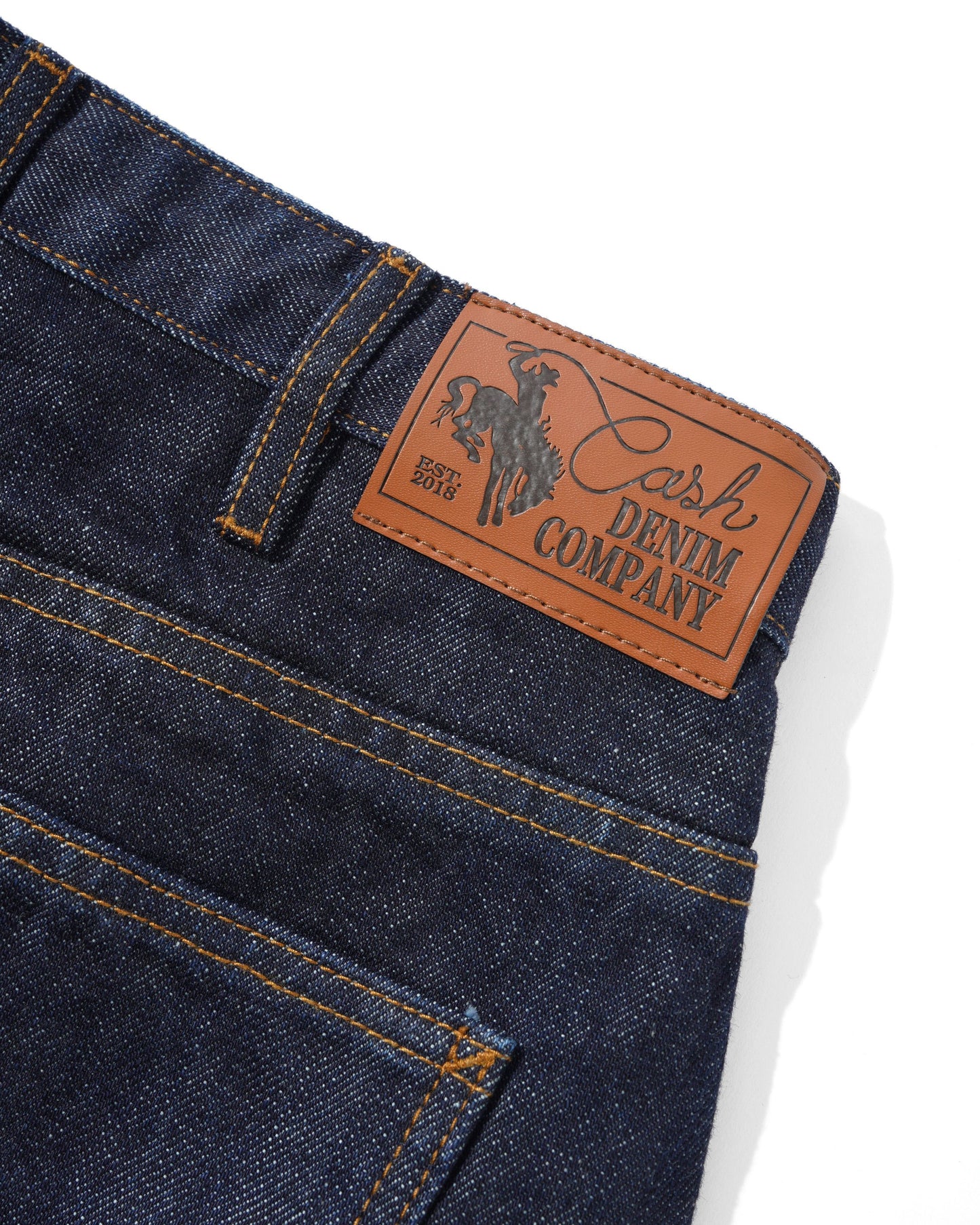 Close-up of dark indigo denim jeans showing the back waistband, belt loop, and a tan leather patch with the Cash Denim Company logo and cowboy graphic.