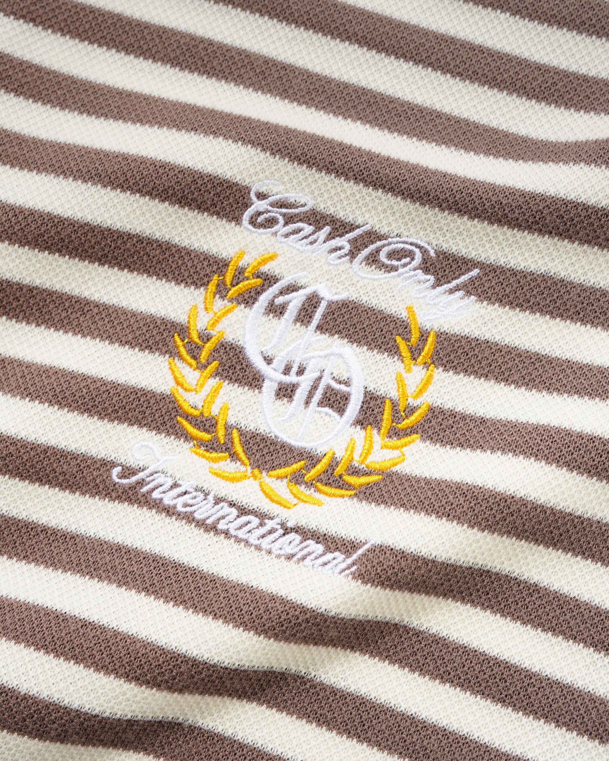 Close-up of a brown and cream striped knit fabric with white and yellow embroidered text reading 'Cash Only International' and a monogram motif.