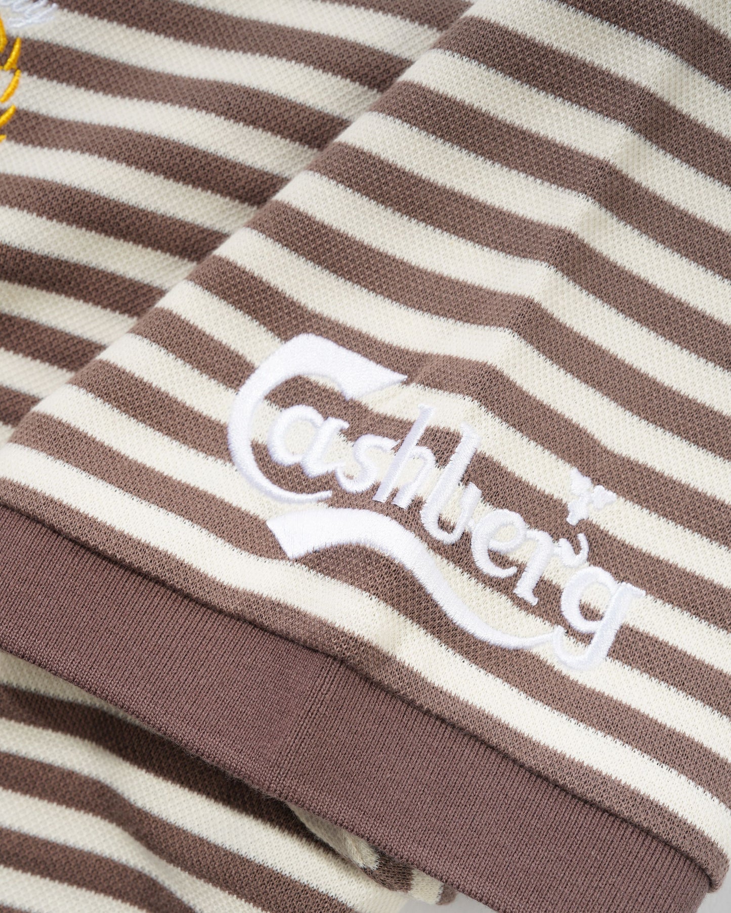 Close-up of a brown and cream striped knit garment with a white embroidered Cashberg logo on the chest.