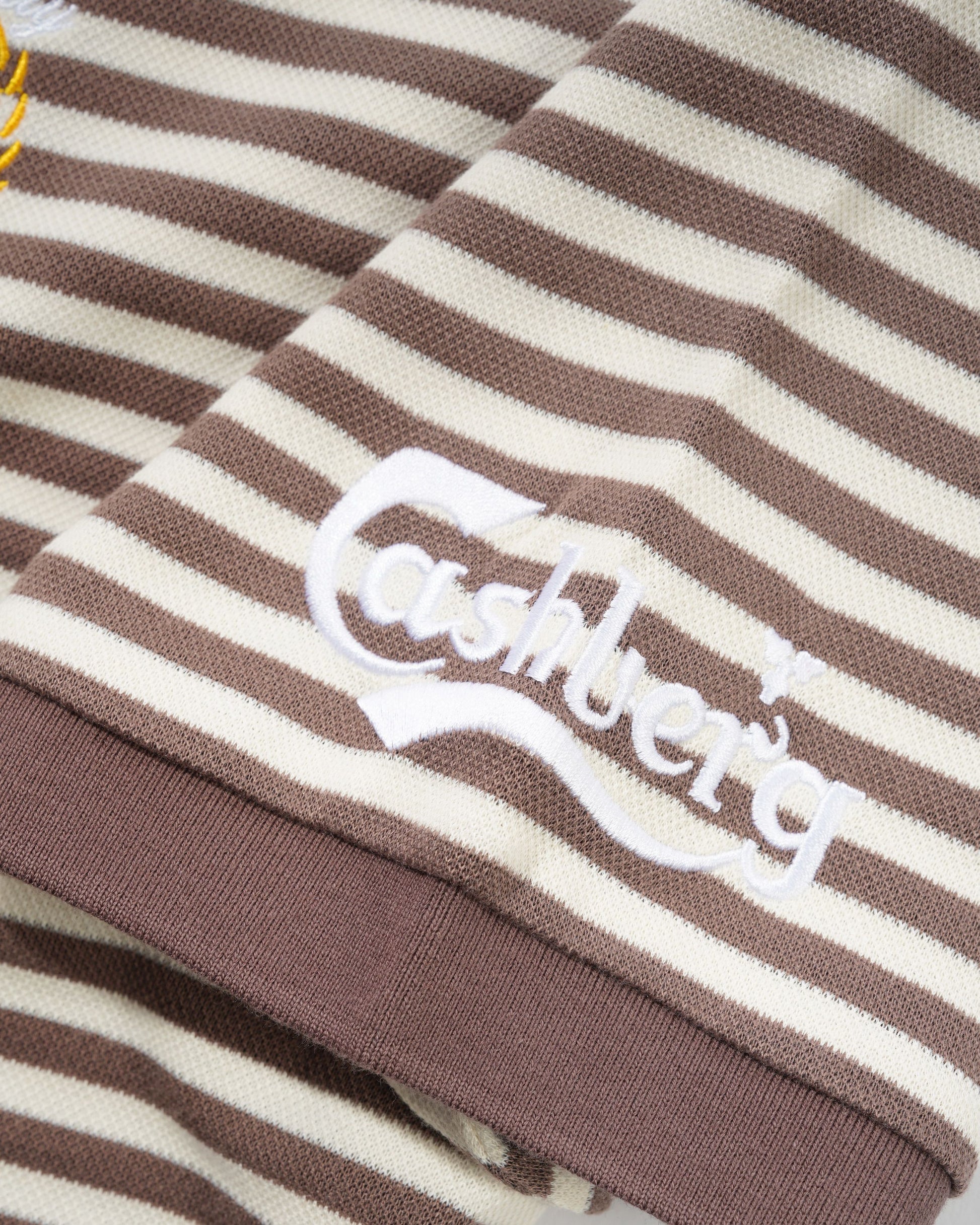 Close-up of a brown and cream striped knit garment with a white embroidered Cashberg logo on the chest.