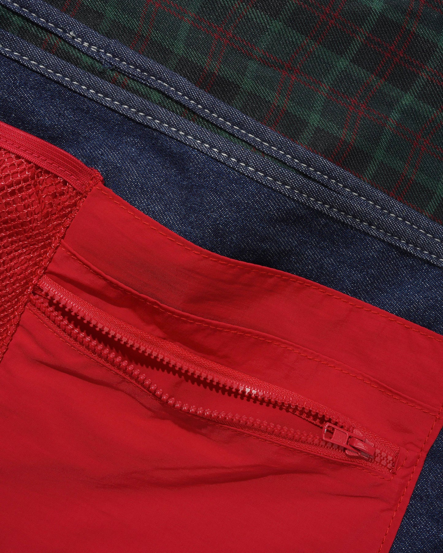 Close-up detail of a jacket showing a red zippered pocket, a red mesh panel, dark blue denim trim, and a green plaid patterned fabric.