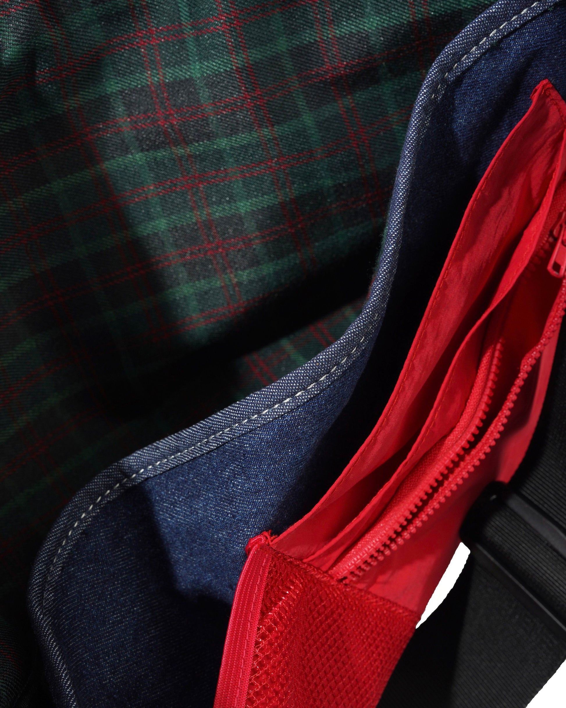 Close-up detail of a bag showing a dark green and red plaid fabric exterior, a dark blue denim-like trim, and a bright red mesh zippered pocket.