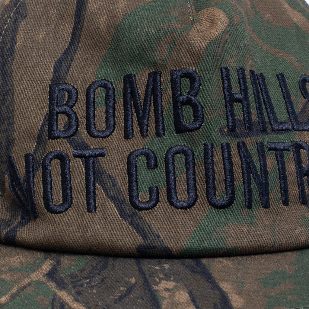 Camouflage cap with 'Bomb Hills Not Country' text on a close-up view