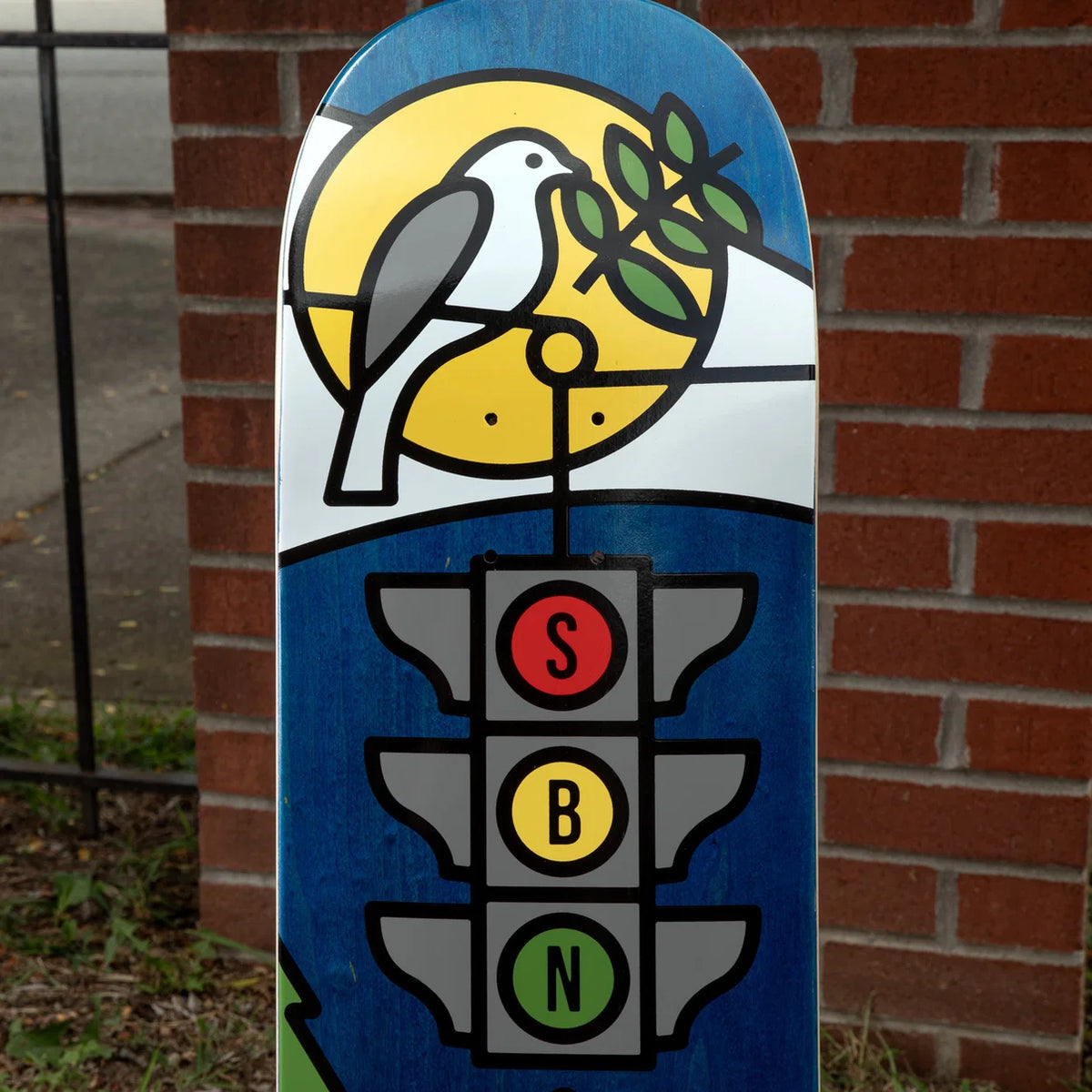 A skateboard deck featuring a graphic design with a white bird perched on a yellow sun and green leaves, above a blue background and a traffic light graphic with red, yellow, and green lights labeled S, B, and N.