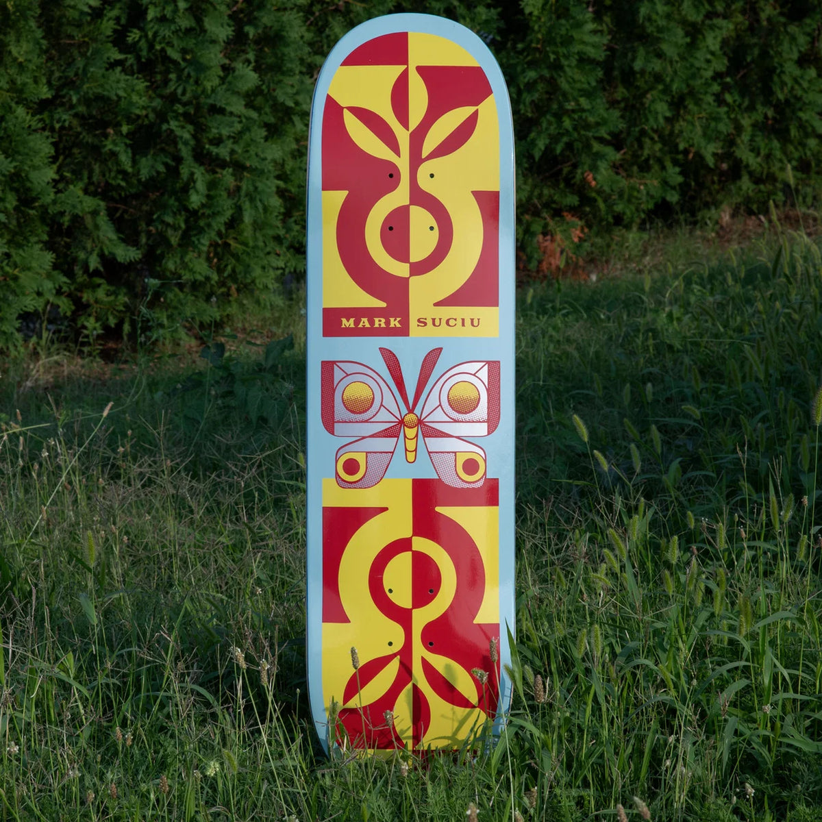 A skateboard deck standing upright in tall green grass, featuring a symmetrical graphic design with red and yellow abstract motifs and a butterfly in the center. The brand name 'MARK SUCIU' is printed in black on the upper section.