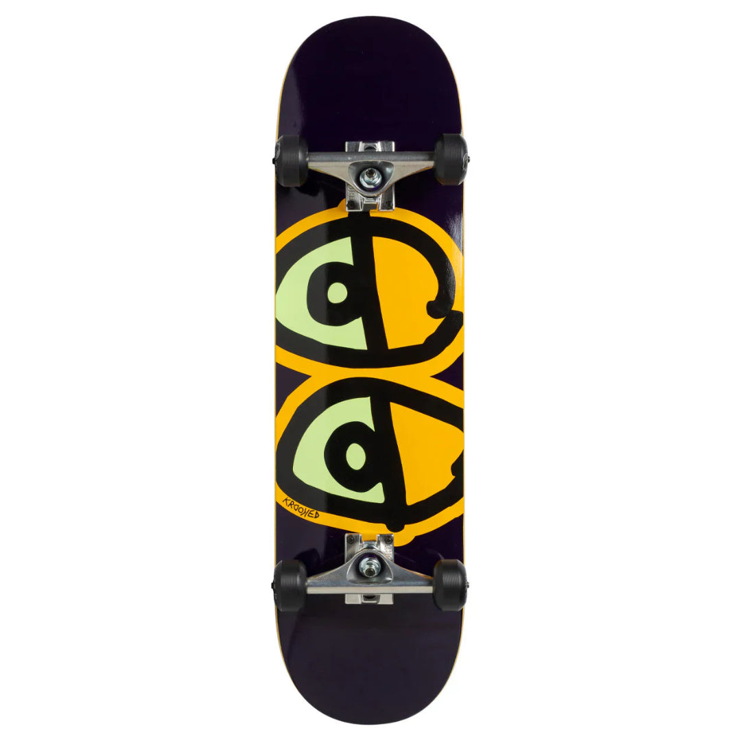 A skateboard shown from the underside, featuring a dark purple deck with a large graphic of two stylized green eyes outlined in black over a yellow background. The skateboard is equipped with silver trucks and black wheels, set against a plain white background.