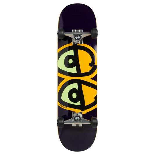 A skateboard shown from the underside, featuring a dark purple deck with a large graphic of two stylized green eyes outlined in black over a yellow background. The skateboard is equipped with silver trucks and black wheels, set against a plain white background.