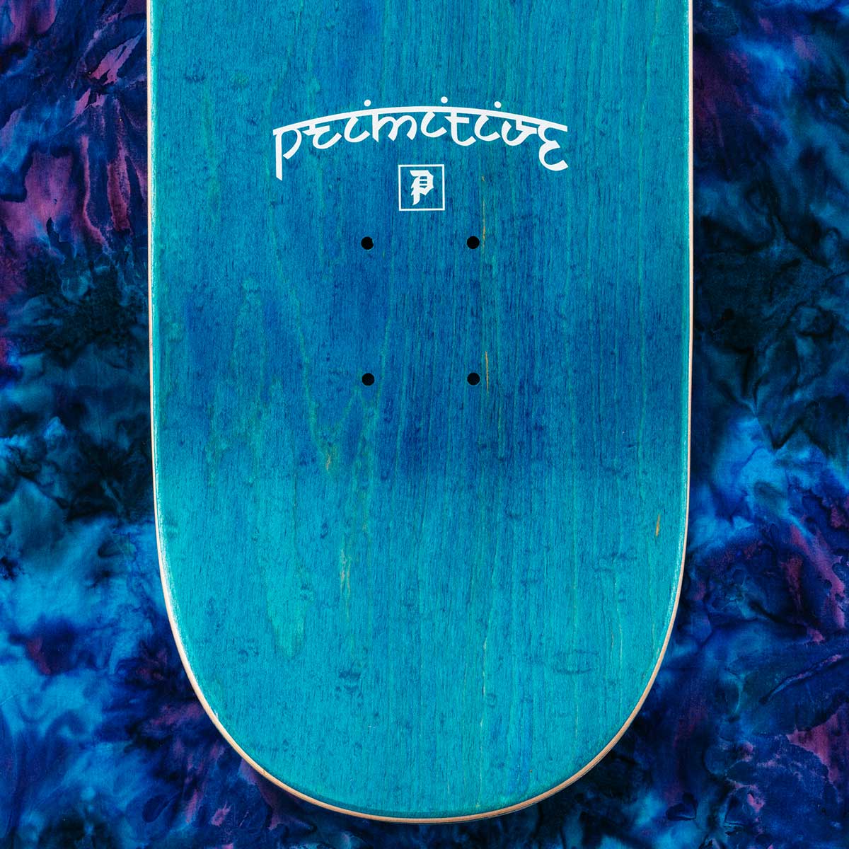 The underside of a skateboard deck with a vibrant turquoise blue wood grain finish, featuring the white 'primitive' brand logo and graphic in the center, set against a dark blue and purple tie-dye background.
