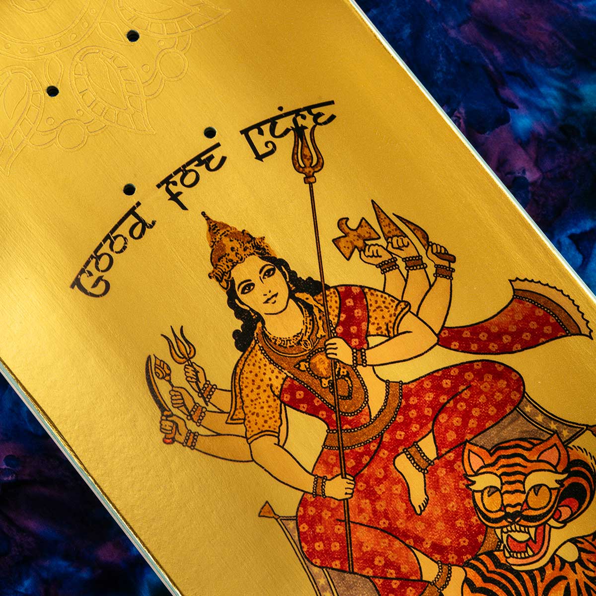 A close-up of a skateboard deck featuring a detailed illustration of the Hindu goddess Lakshmi on a metallic gold background, with Devanagari text and a tiger at the bottom.