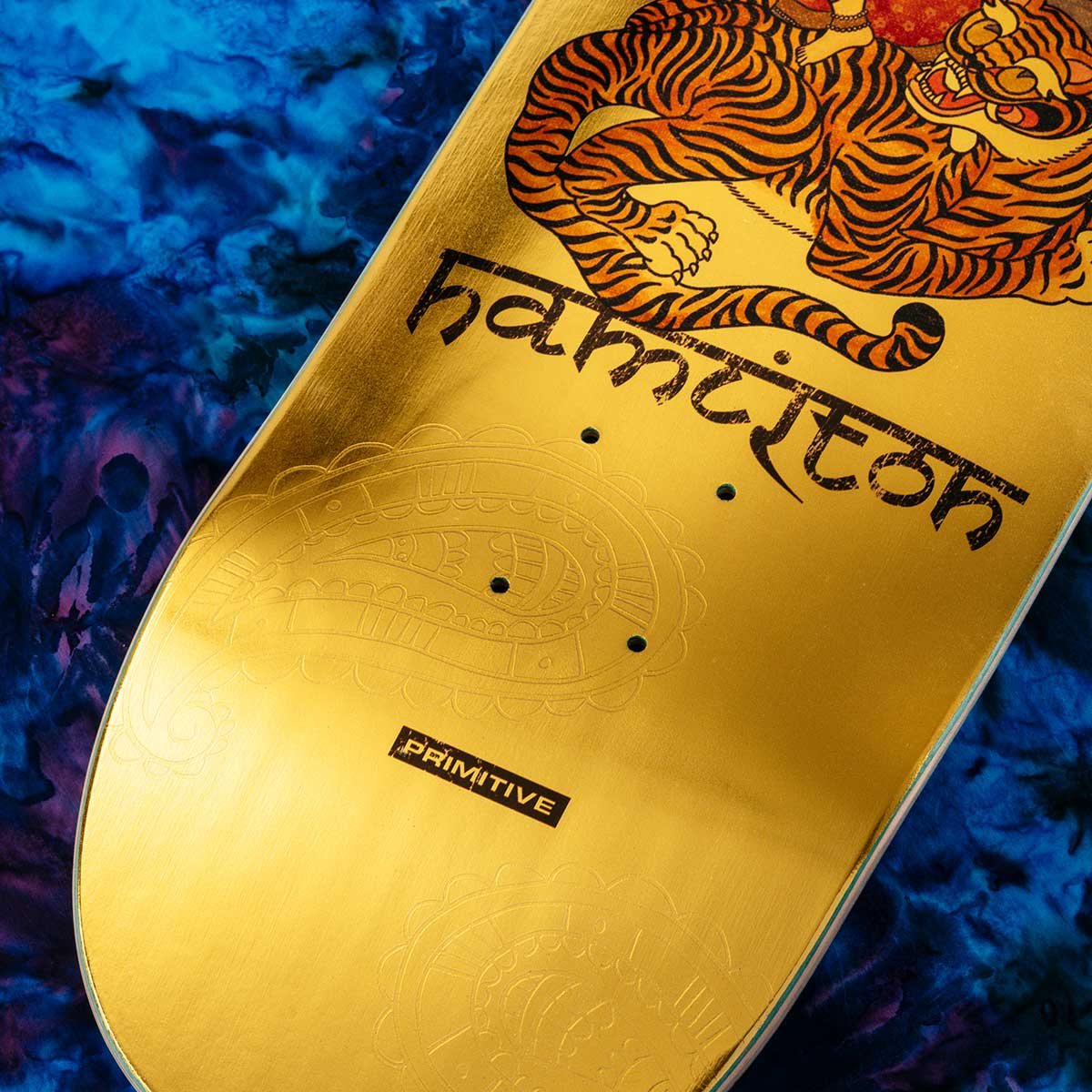 A close-up of a skateboard deck featuring a metallic gold base with a large orange tiger graphic, the word 'hamiton' in stylized black lettering, and a small 'PRIMITIVE' brand label near the bottom, set against a blue marbled background.