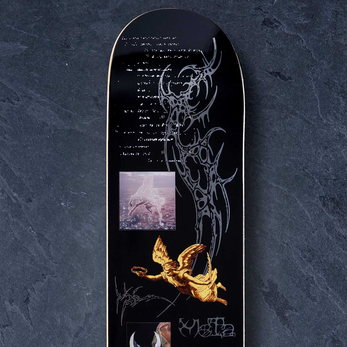 A black skateboard deck featuring a large silver tribal wing design, a golden angel illustration, white script text, and a small square photo inset, photographed against a dark textured background.