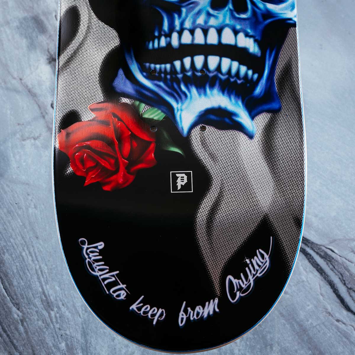 A close-up of a skateboard deck featuring a graphic design with a blue skull, a red rose, and white script text reading 'Laugh to keep from crying' on a black background.