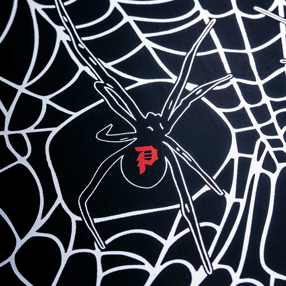 A close-up of a black fabric surface printed with a white spiderweb pattern and a stylized spider graphic in the center, featuring a small red stylized 'P' emblem.