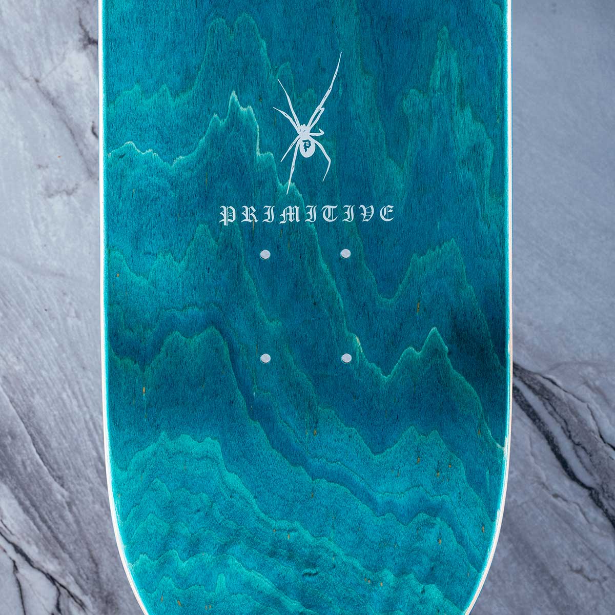 A close-up of a skateboard deck underside featuring a teal and turquoise marbled wood grain pattern with a white spider graphic and the word 'PRIMATE' in gothic lettering.