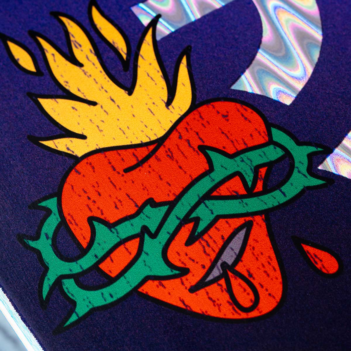 A close-up of a graphic design featuring a red heart pierced by a green thorn crown, with a yellow flame rising from the top, set against a dark navy background with a holographic foil reflection in the upper right.