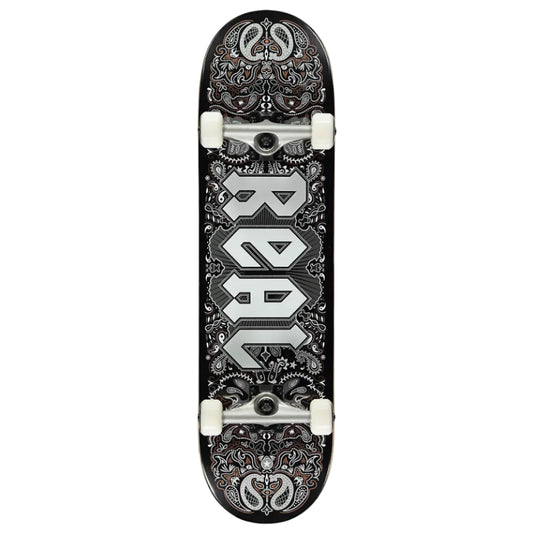 A skateboard deck shown vertically against a light gray background, featuring a black base with intricate silver and white paisley-style graphics and the word 'REAL' printed vertically in large silver letters.