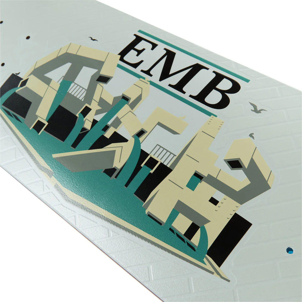 A close-up of a skateboard deck featuring a stylized graphic of the word 'EMBASSY' in bold black letters with teal and beige geometric letterforms, on a white deck with a subtle diamond pattern.