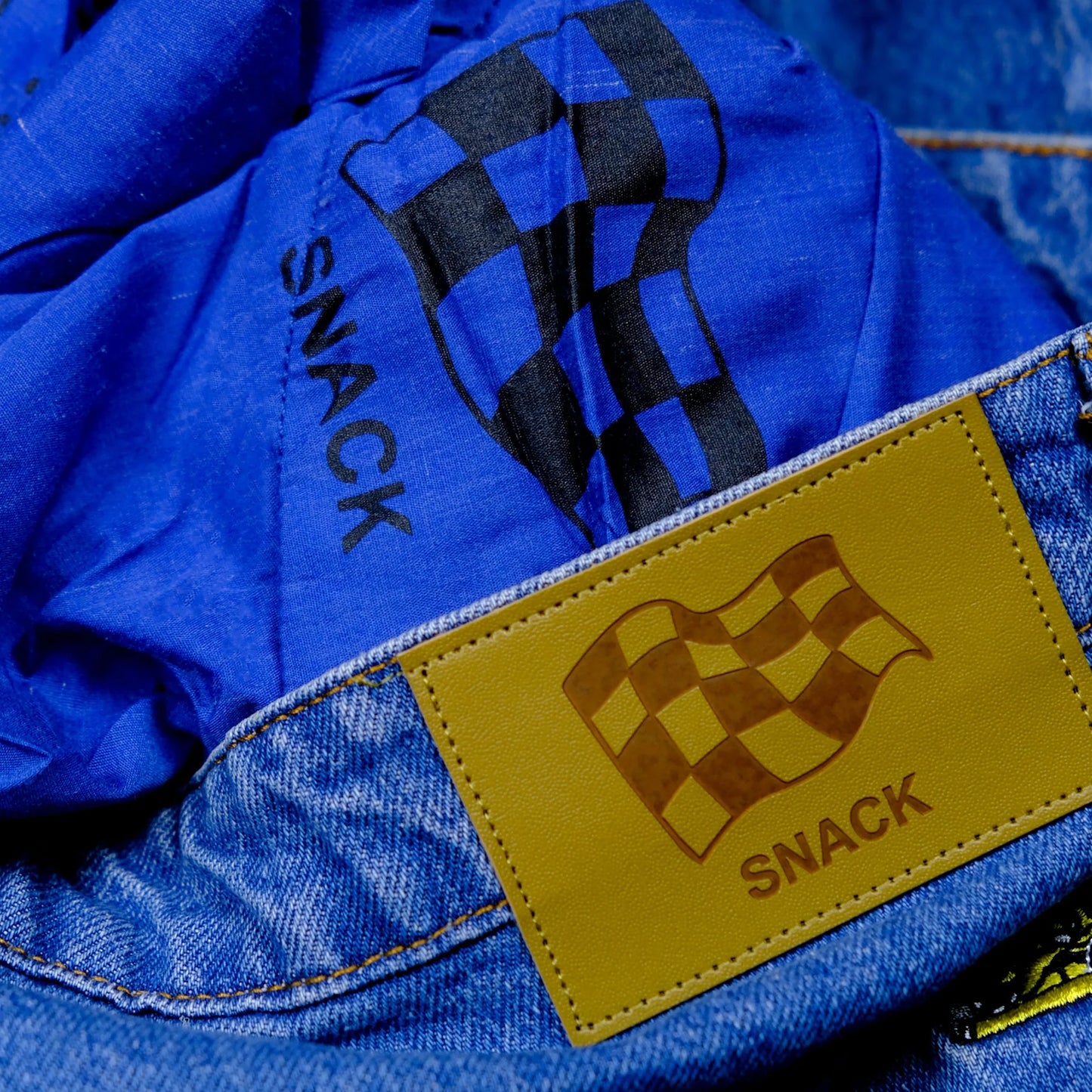 Close-up of a blue jacket with a yellow patch featuring a checkered flag design and the word 'SNACK'.