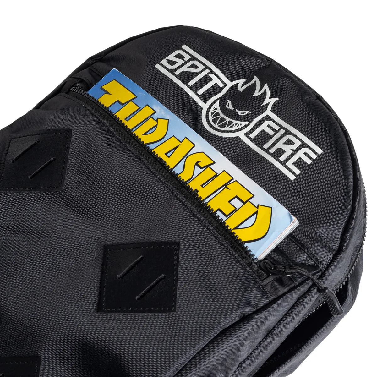 A close-up of a black backpack featuring a prominent white 'SPIT FIRE' logo with a black skull graphic on the top, and a horizontal 'THRASHER' patch in yellow and blue sliding over the front zipper pocket.