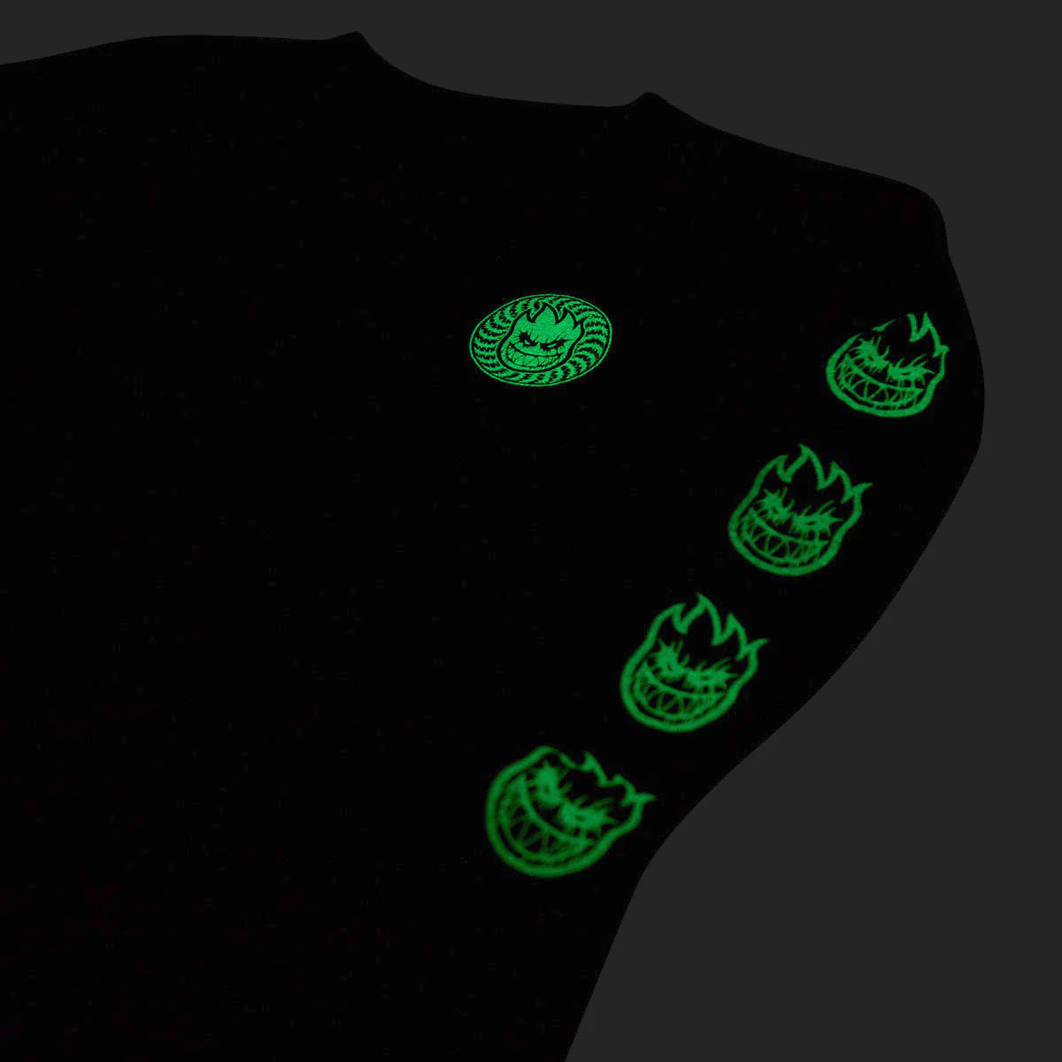 A black garment, likely a sweatshirt, shown on a dark background with glowing green graphic prints. The design features a circular skull-like emblem on the upper chest and four flame-shaped skull graphics running down the sleeve.