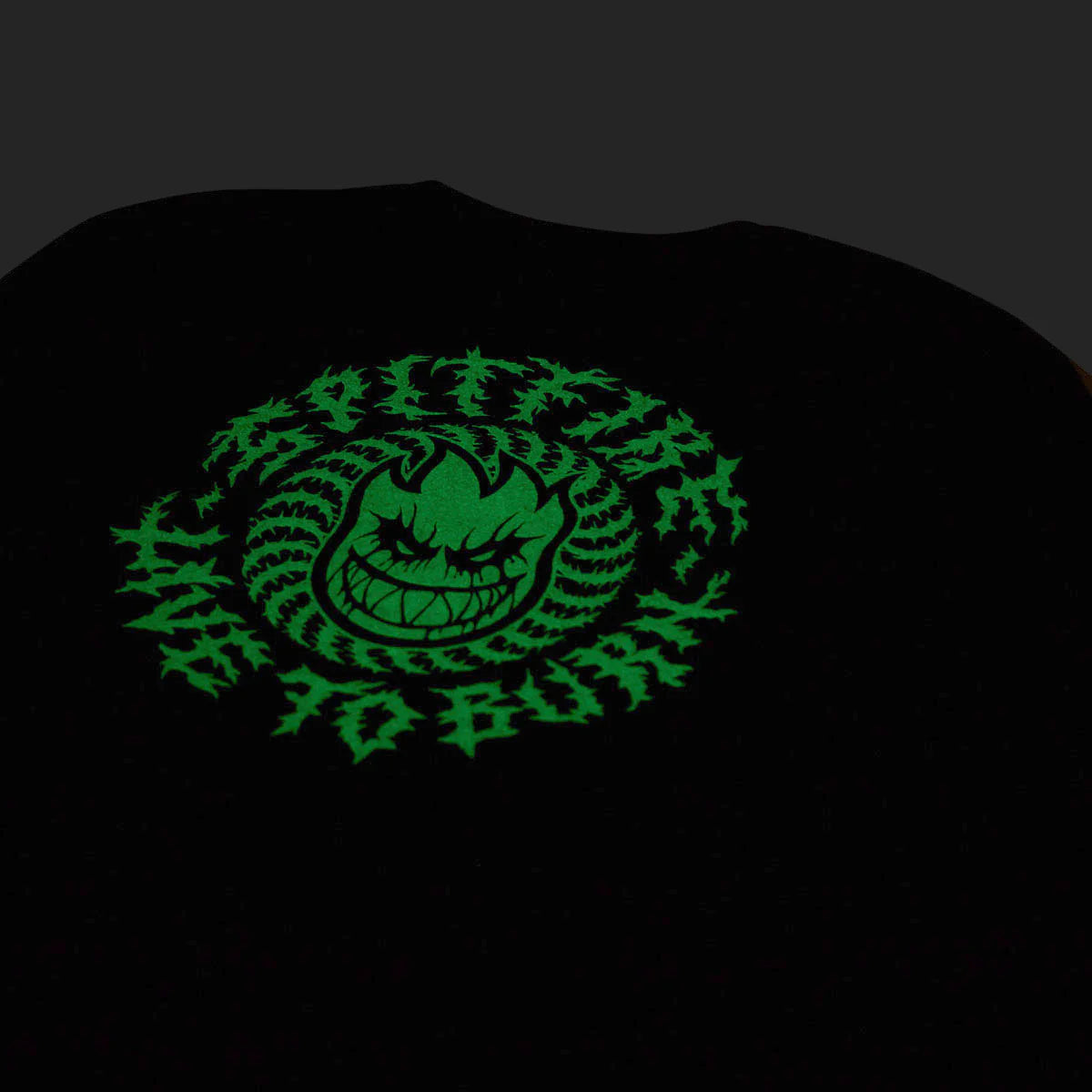 A black t-shirt featuring a glowing green graphic of a demonic face surrounded by sharp thorn-like patterns, shown against a dark background to highlight its glow-in-the-dark effect.
