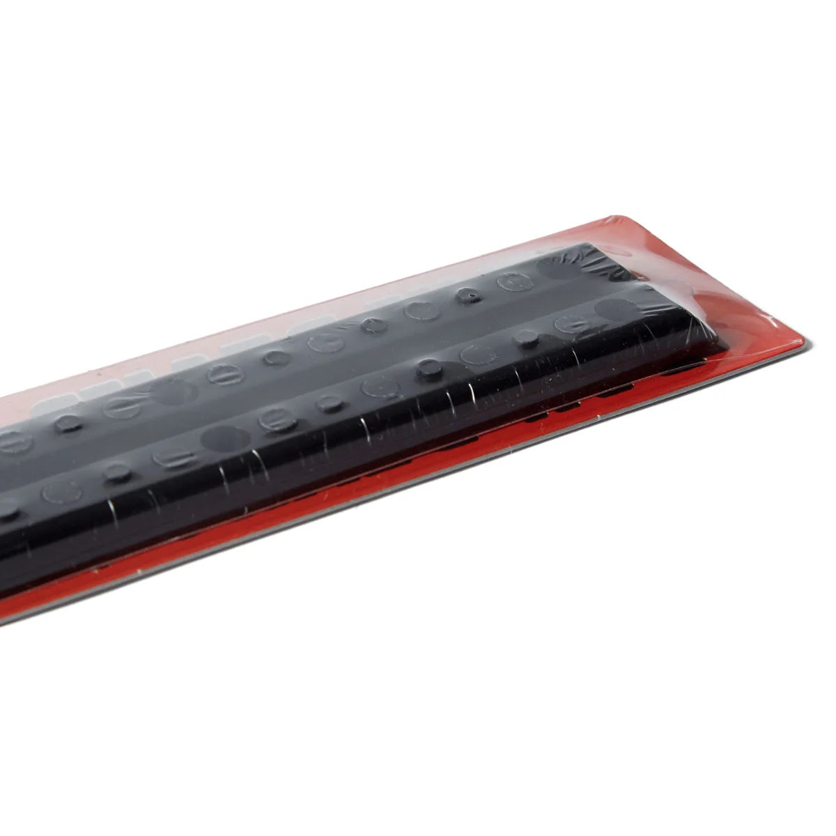 A close-up of a black textured strip with circular raised dots, wrapped in clear plastic and set on a red backing, shown against a plain white background.
