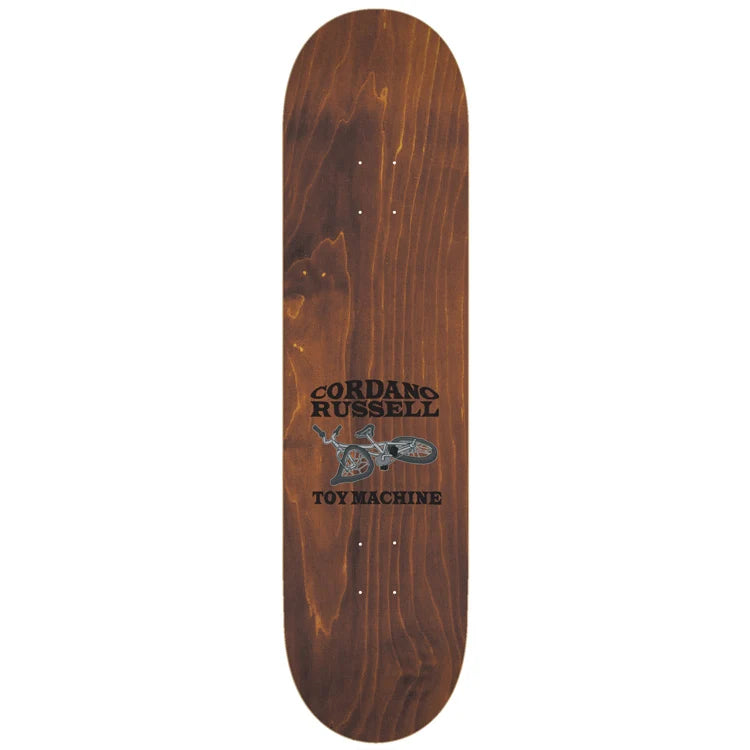 A wooden skateboard deck with a warm brown wood grain finish, featuring the 'Cordano Russell Toy Machine' logo printed in black and gray in the center, shown against a plain white background.