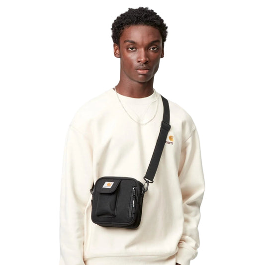 Man wearing a white sweatshirt and dark jeans with a black crossbody bag on a light gray background